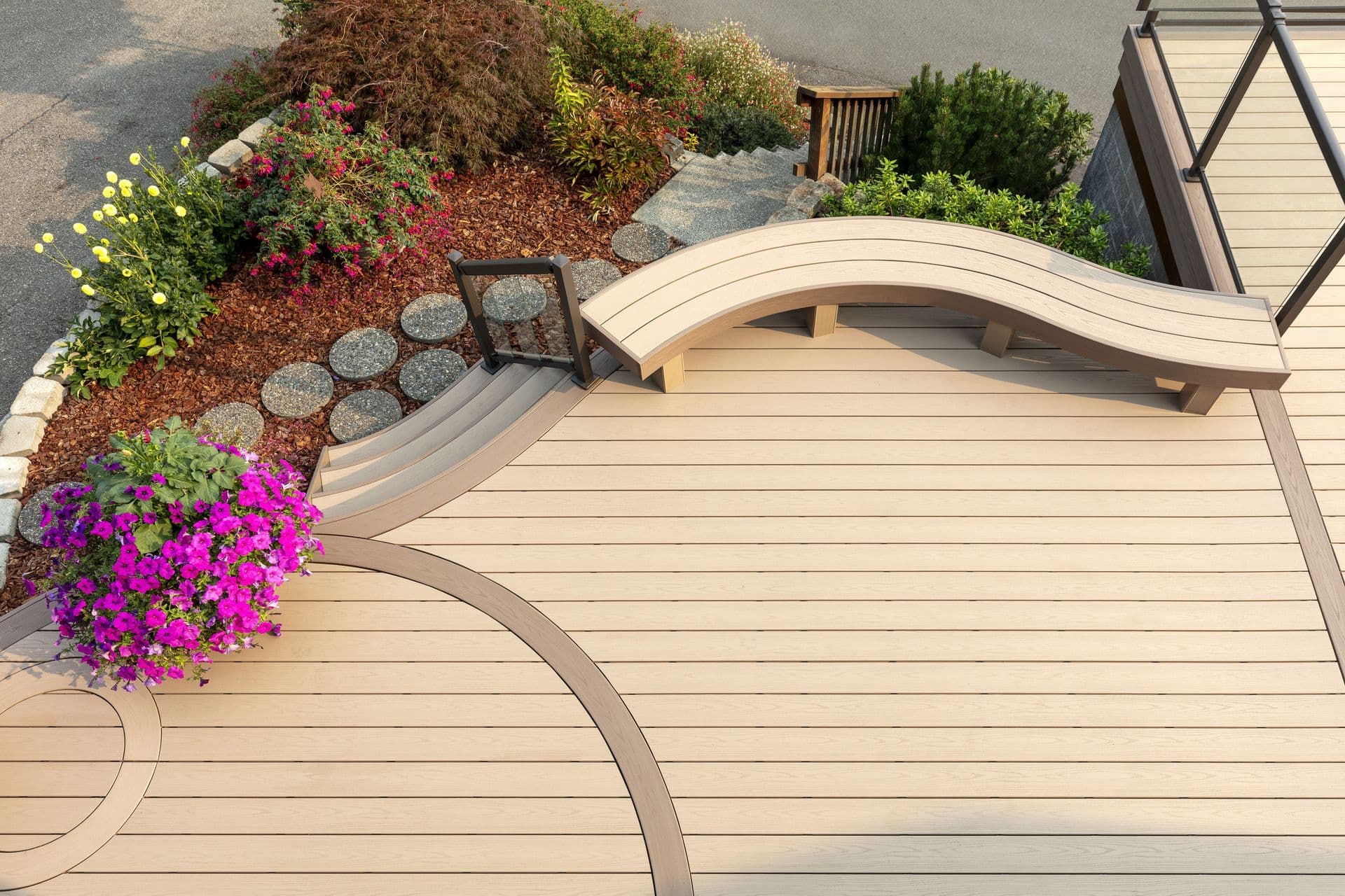 Curved composite deck with light tan boards, featuring a built-in curved bench, decorative inlay pattern, and steps leading to a landscaped garden with flowers and stone pavers.