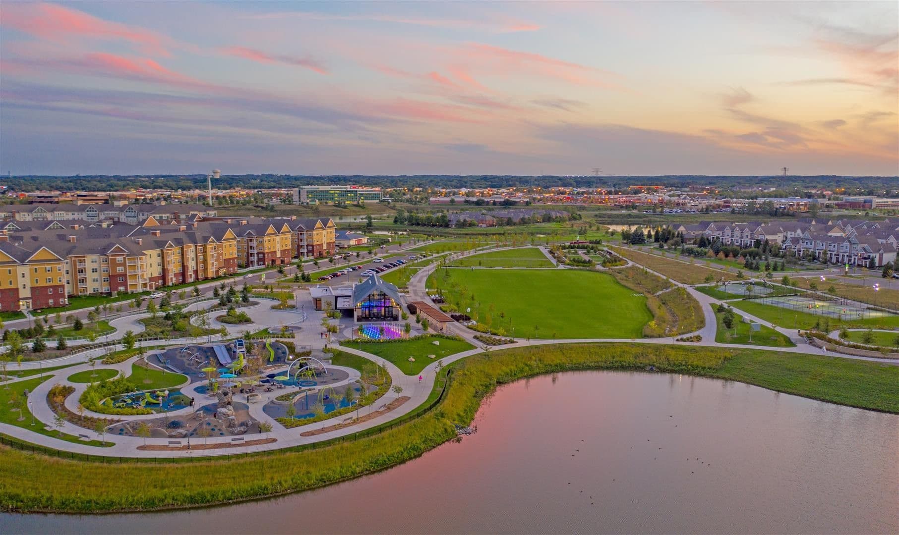 Aerial view of Maple Grove, Minnesota, showing a park, pond, walking paths, nearby apartments, and neighborhoods at sunset.