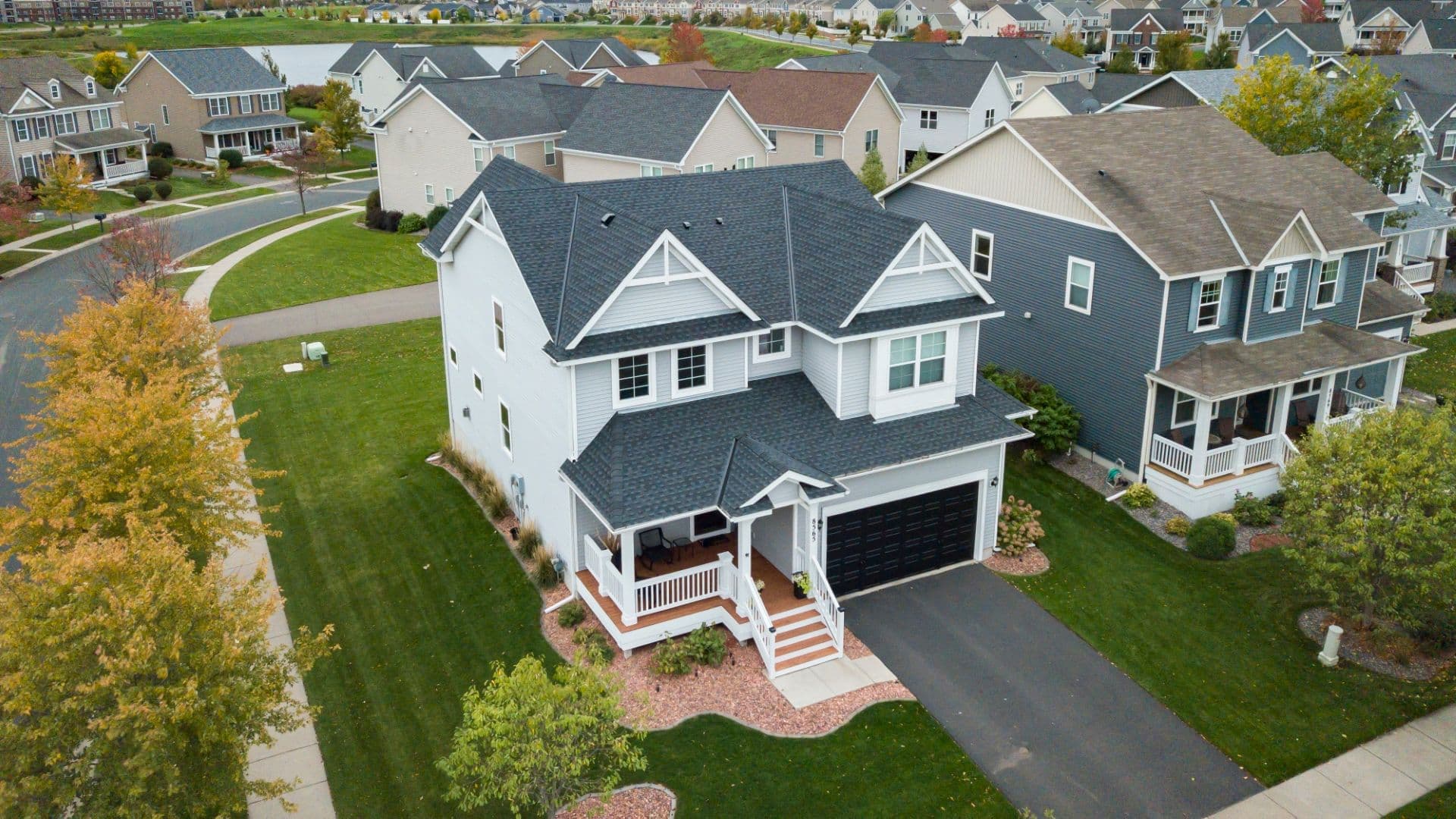 An aerial view of a light gray house with new dark gray shingle roof and a corner front porch and deck.