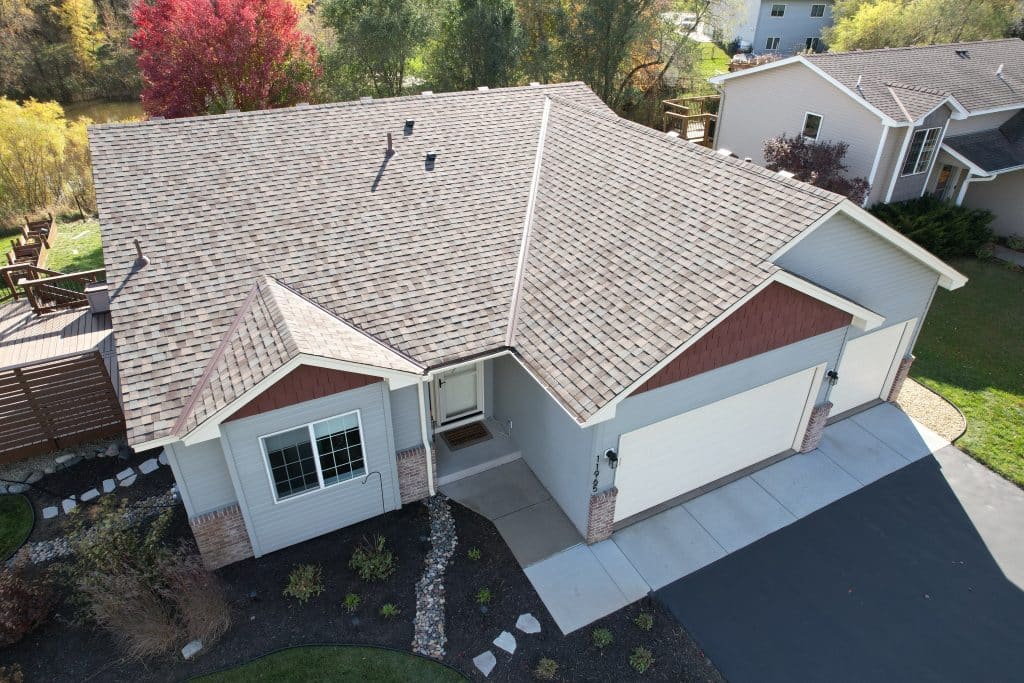 Aerial view of a house with a new roof and an attached garage, featuring a well-maintained backyard deck with a grill and seating area, surrounded by green lawn and landscaping.