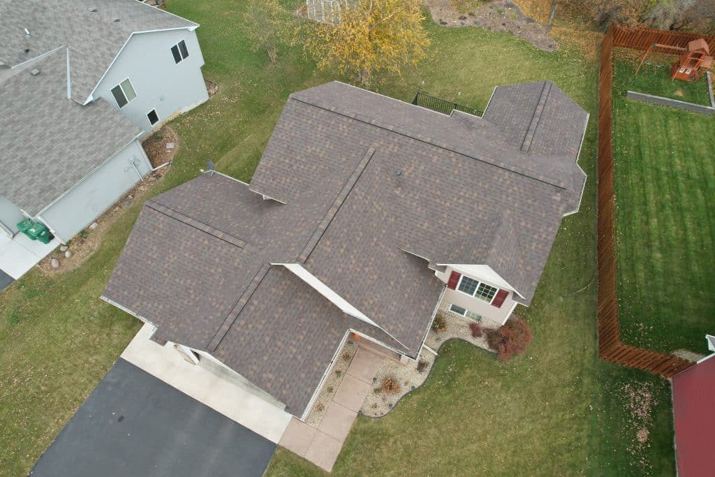 Aerial view of a house with a newly installed roof, situated in a spacious yard with autumn foliage in the background, highlighting the home's clean driveway and landscaped front yard.