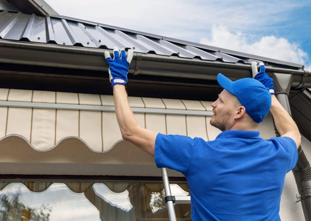 A worker in blue uniform and gloves installs a metal gutter system on a house, standing on a ladder, with a clear blue sky in the background.